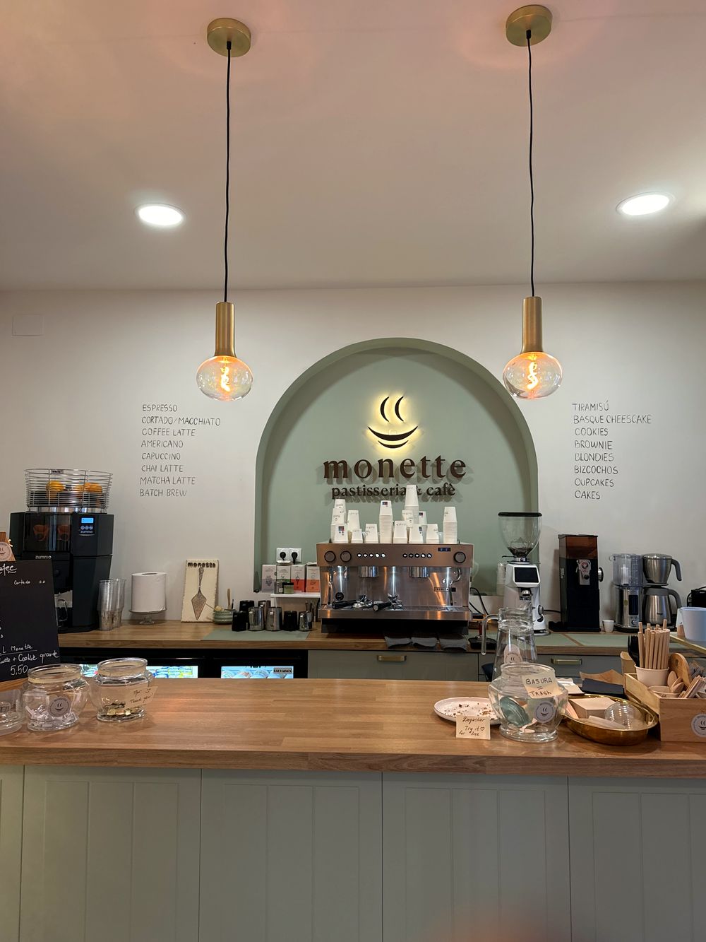 El mostrador de Monette: nicho en arco verde salvia con el logo iluminado, mostrador de roble y lámparas de latón